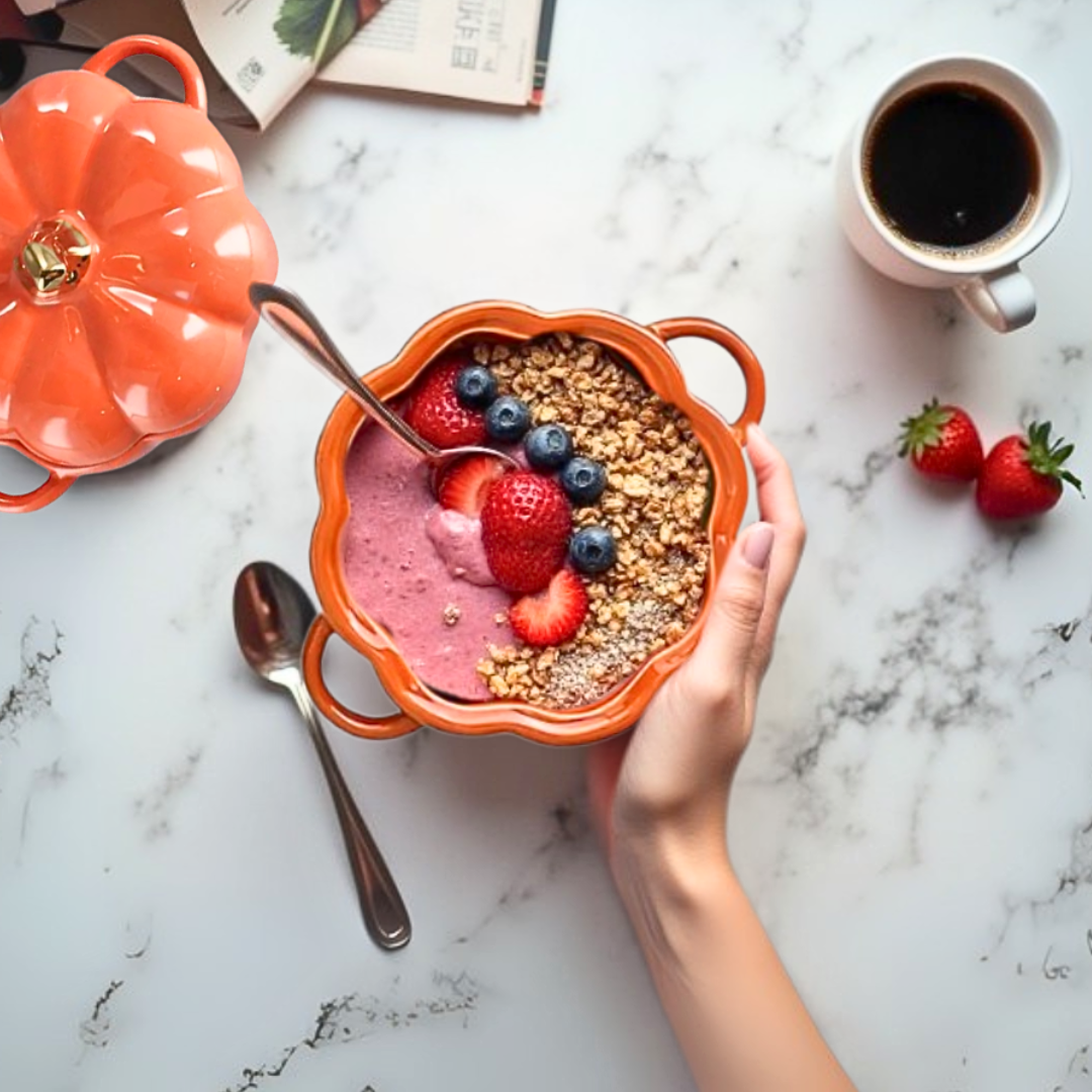 Orange pumpkin-shaped ceramic bowl with granola and berries on a marble surface with coffee and strawberries.