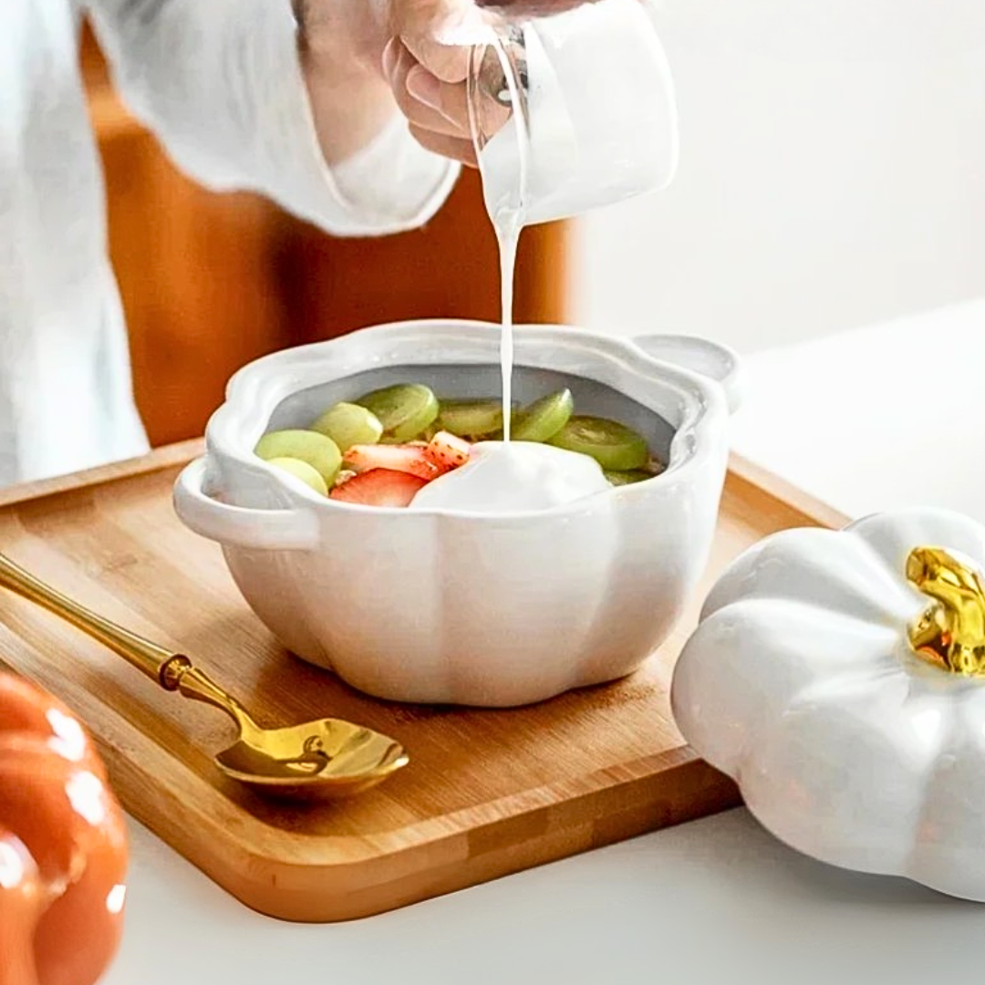 White ceramic pumpkin-shaped bowl with fruit on a wooden tray, being poured into by a person.