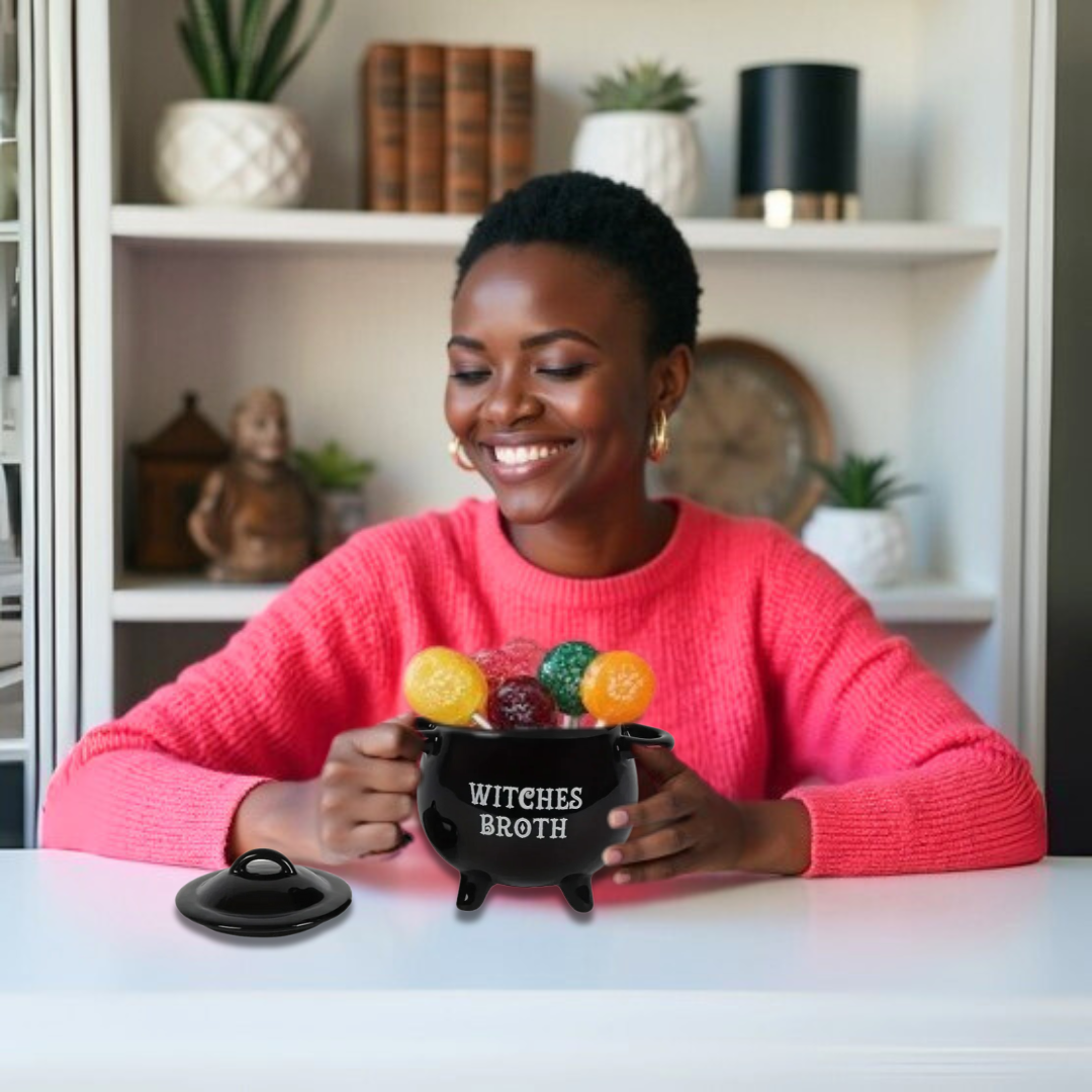 Woman holding a black ceramic cauldron mug labeled 'Witches Broth' with colorful gumballs, smiling in a cozy room.