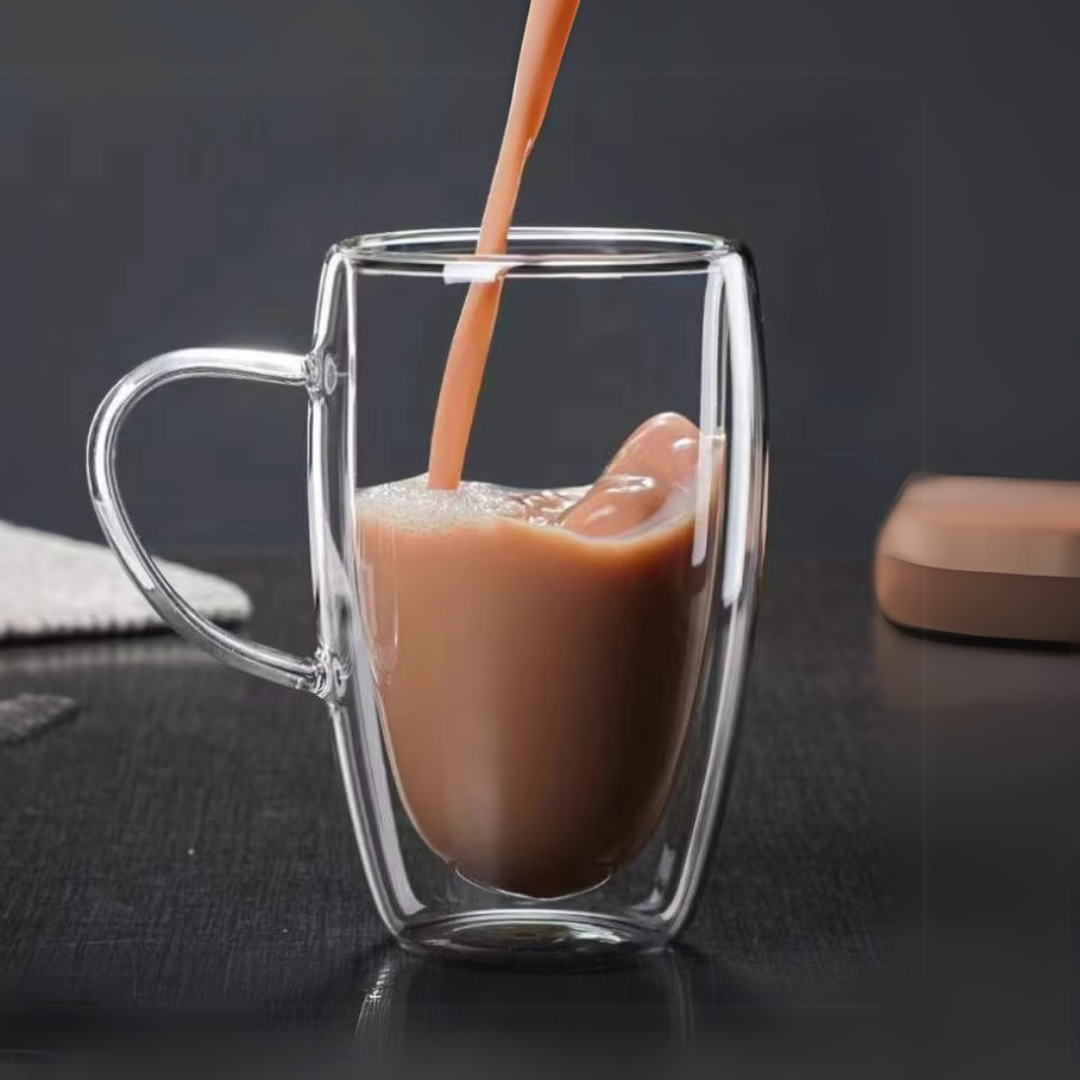 Glass mug with a double wall design being filled with a creamy hot chocolate on a dark table.