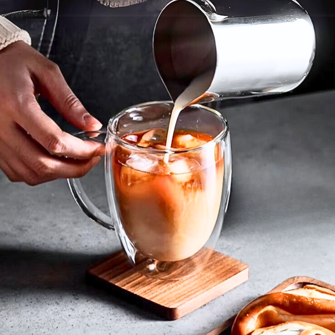 Person pouring milk into a glass of iced coffee on a wooden coaster.