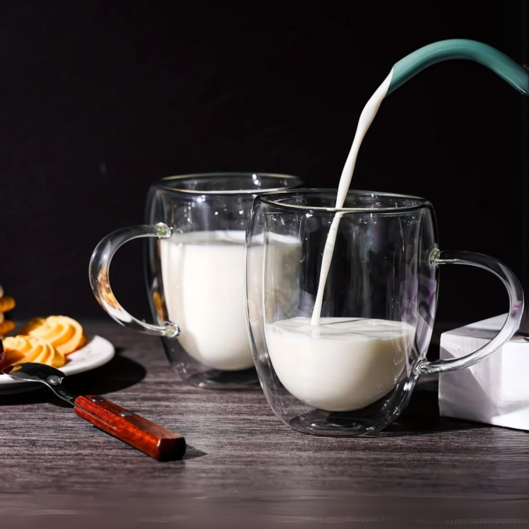 Glass mugs with handles on a wooden table, one being filled with milk