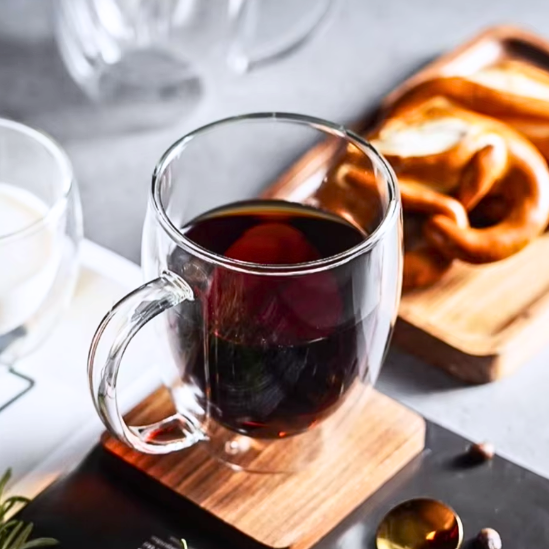 Clear glass mug with coffee on a wooden coaster, with bread in the background