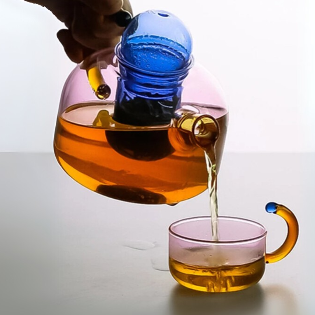 Tea being poured from a clear glass teapot with a blue tea ball into a matching glass cup