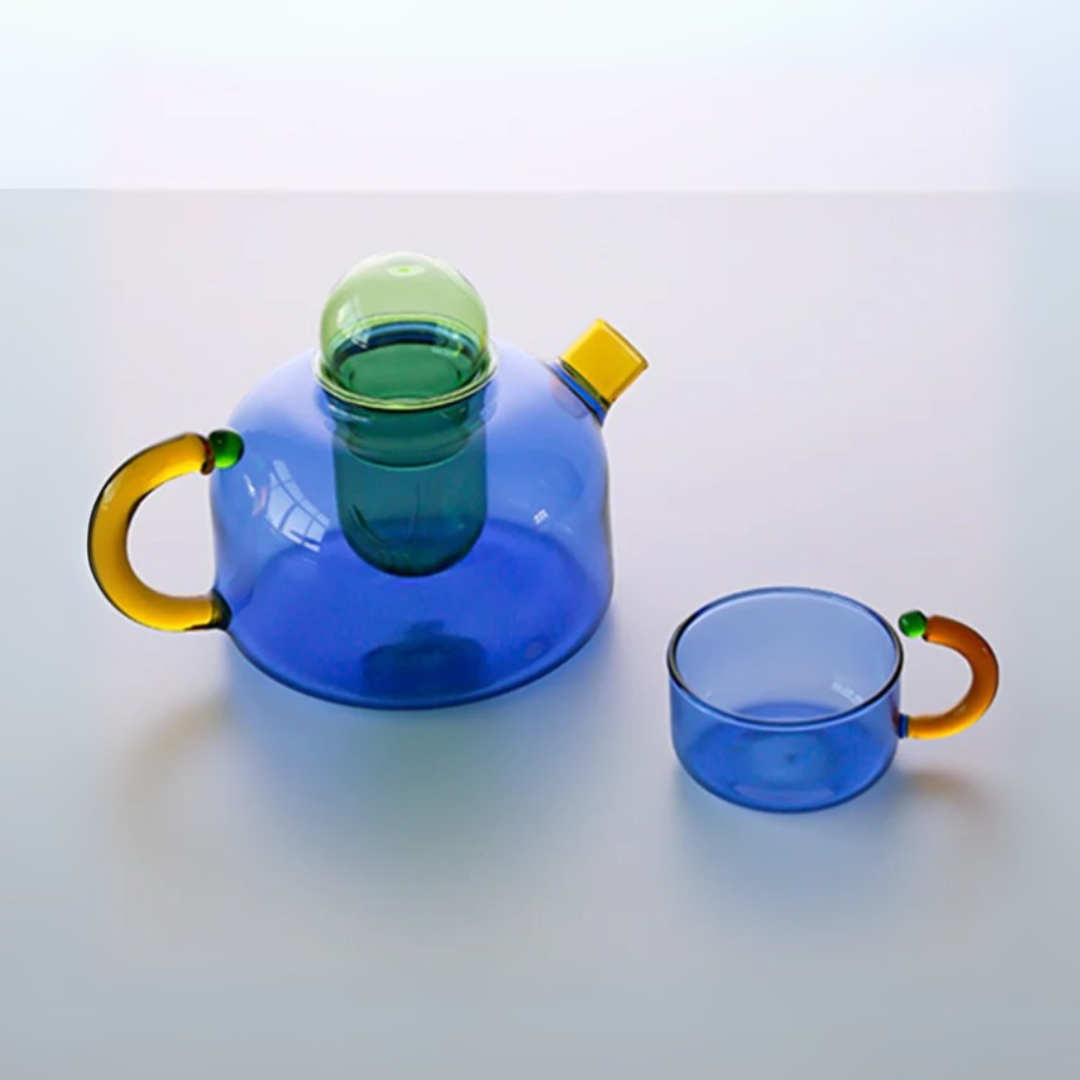 Blue glass teapot with yellow handle and green lid, accompanied by a matching blue glass cup with yellow handle