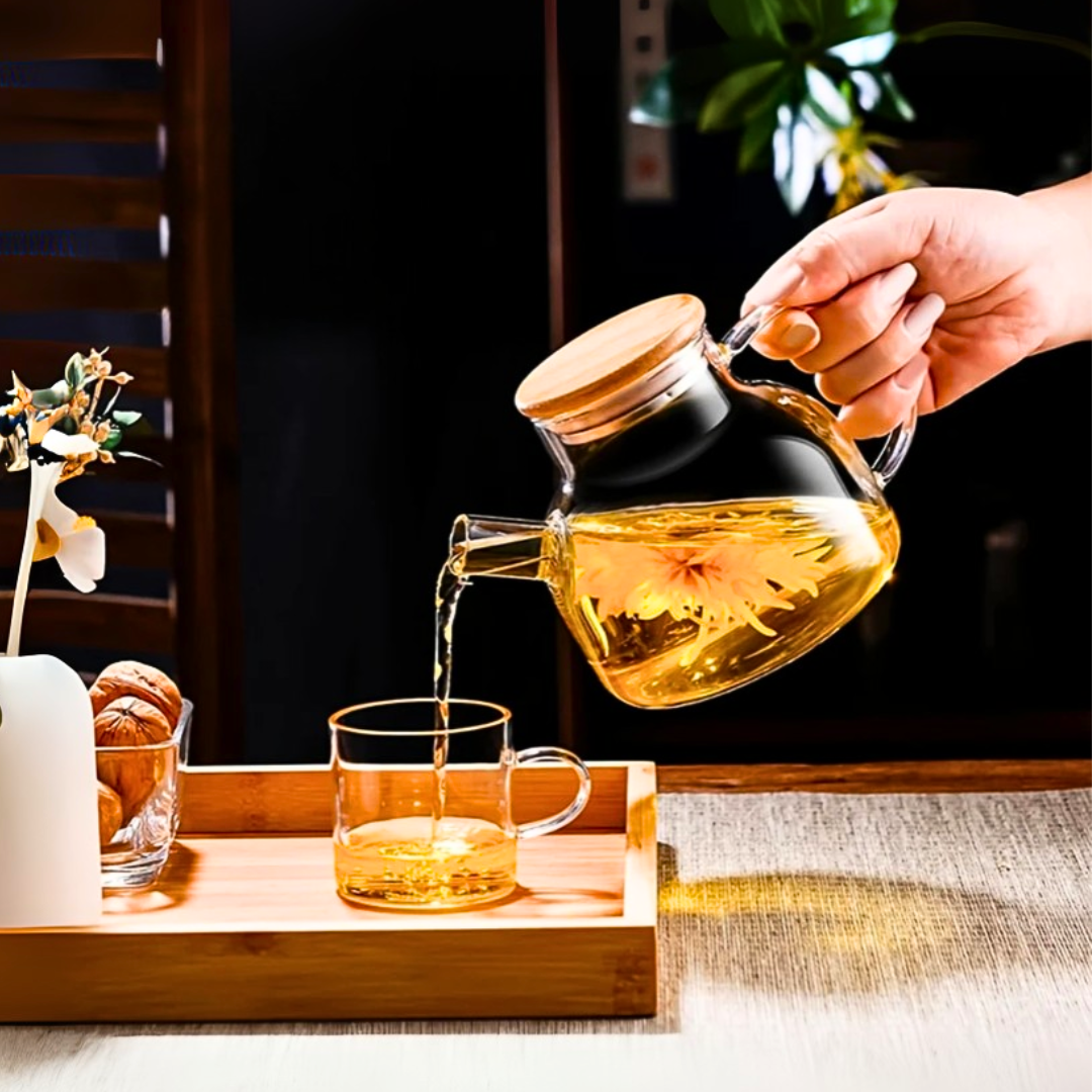 Hand pouring tea from a glass kettle into a glass on a wooden tray.