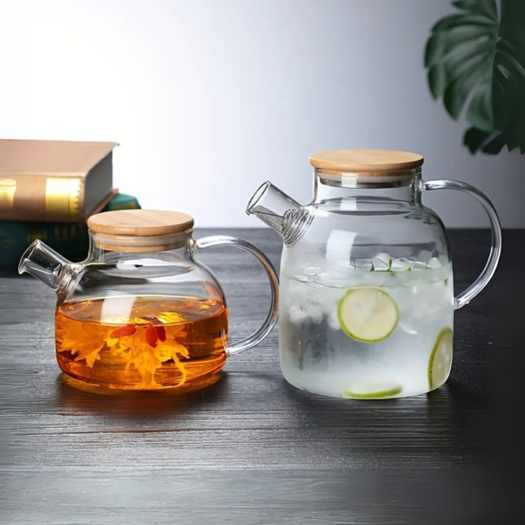 Two glass kettles on a wooden table, one with a wooden lid containing a hot beverage and the other with a wooden lid and a clear lid containing a cold drink.
