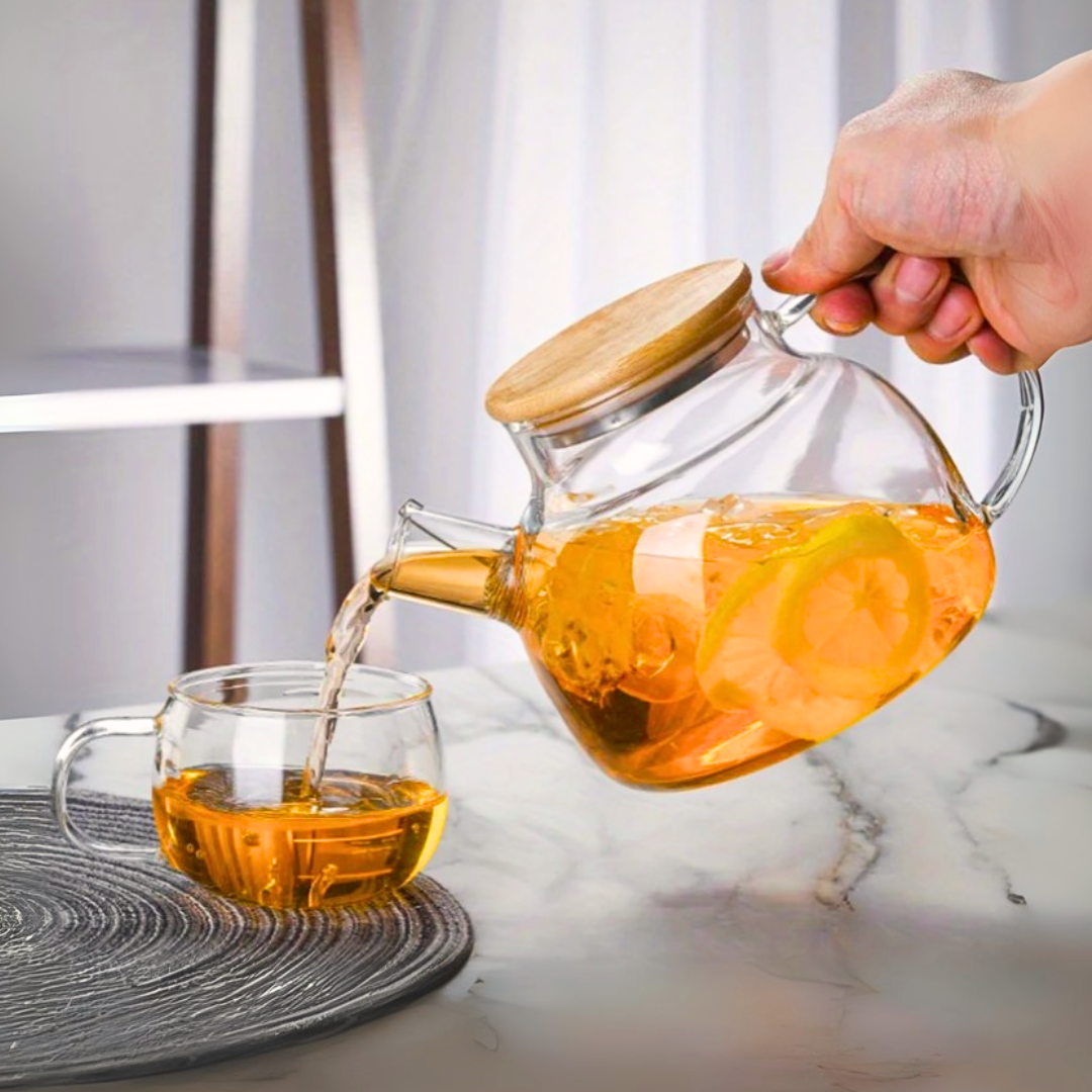 Glass kettle with wooden lid being filled with tea into a glass mug on a marble counter.
