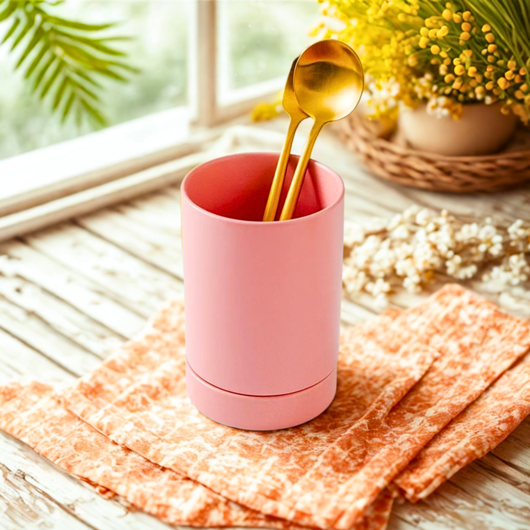 Pink ceramic utensil crock with gold spoons on a wooden table with a window and plants in the bright kitchen.