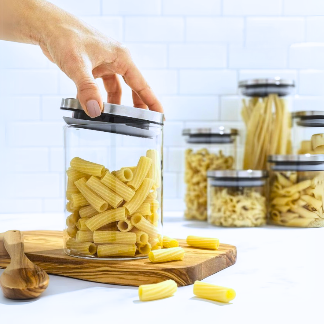 Hand reaching into a glass jar filled with pasta on a wooden board, with more jars of pasta in the background.