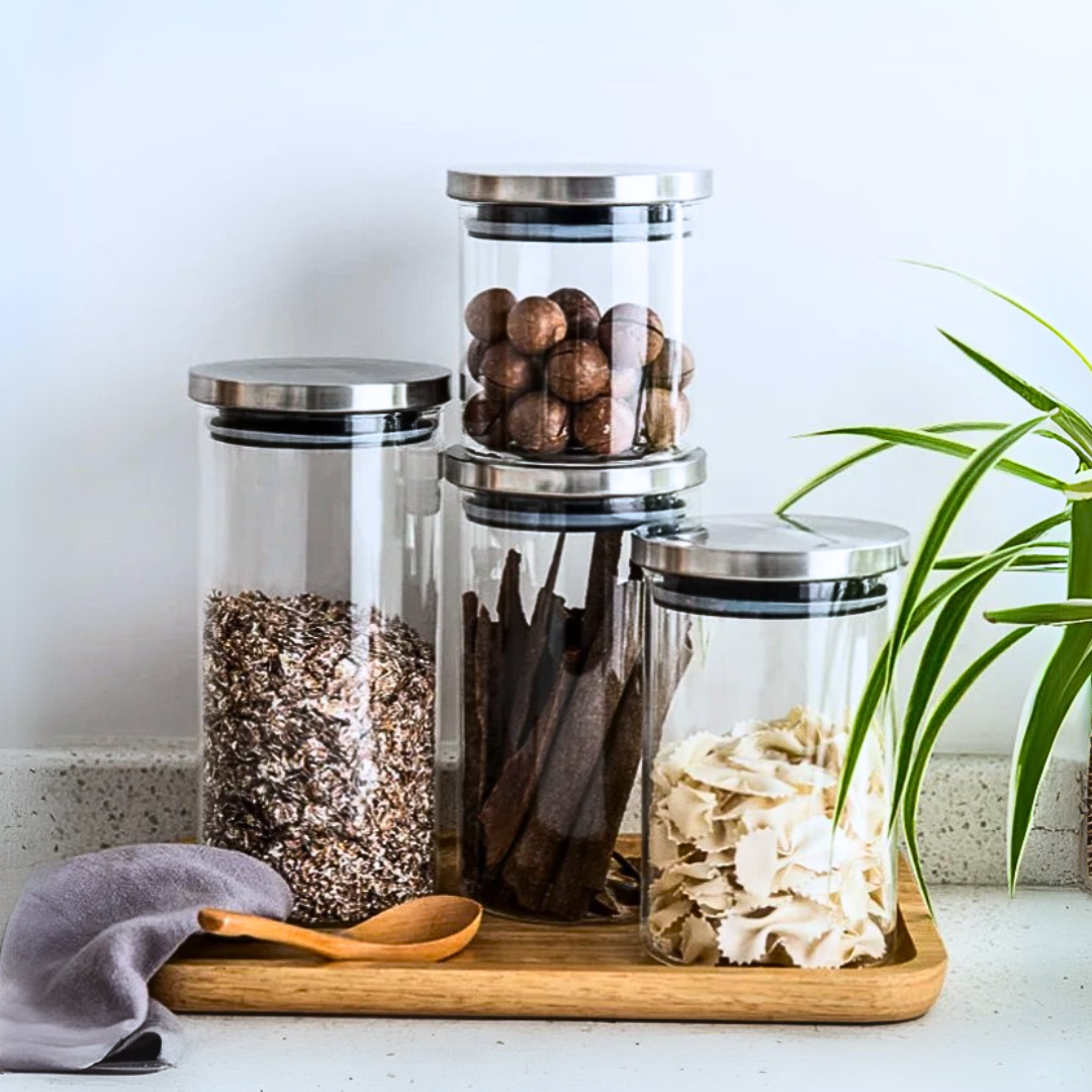Set of glass storage jars with airtight stainless steel lids on a wooden tray, containing various items, with a plant in the kitchen.